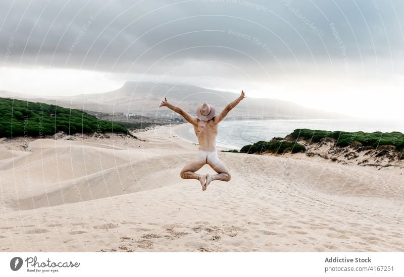 Anonymous sporty naked man jumping on sandy desolate seacoast nude active carefree freedom beach scenic travel seashore hat fit excited bare unclothed enjoy