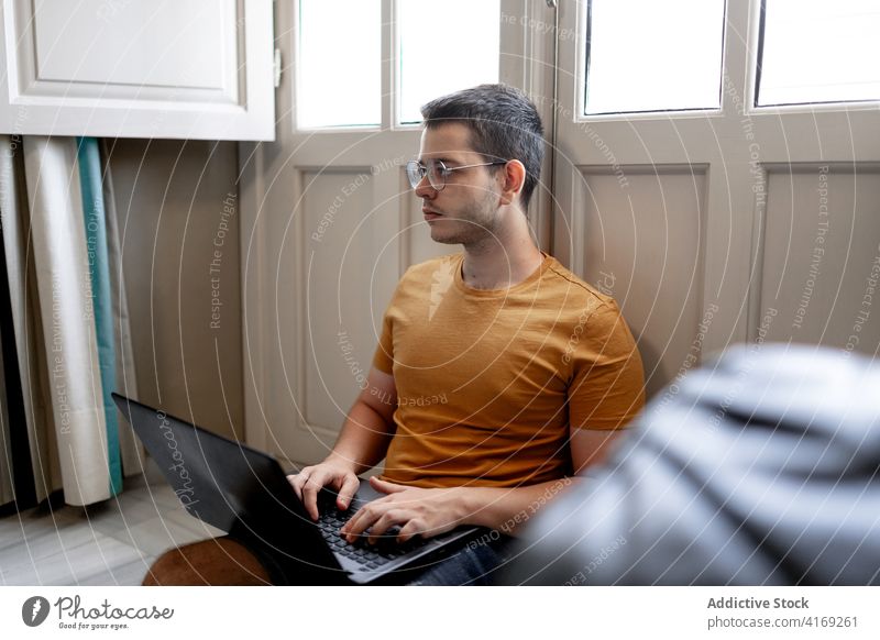Busy man working on laptop in living room freelance remote home typing project concentrate browsing male casual outfit floor sit window legs crossed device job