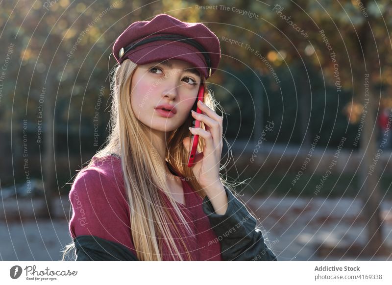 Stylish young woman talking on phone in autumn park smartphone trendy fashion colorful red millennial female hat device gadget style connection modern mobile