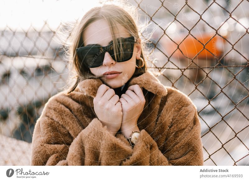 Stylish woman in warm jacket in city fur style trendy outfit determine sunglasses outerwear female net fence street urban cool fashion serious town calm