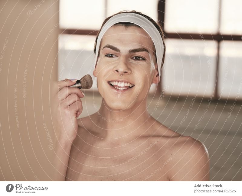 Shirtless young guy applying makeup man androgynous studio foundation brush transgender shirtless mirror male queer cheek cosmetic headband prepare skin care