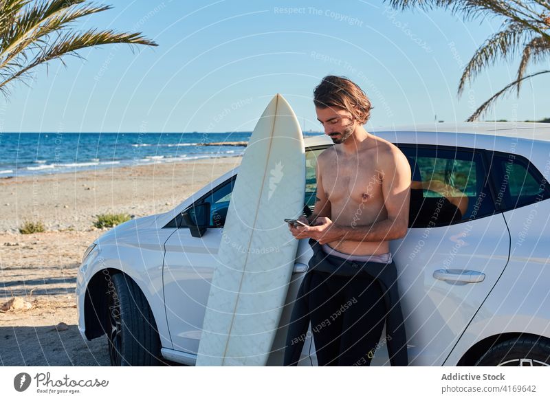 Shirtless man with surfboard near car using smartphone surfer browsing beach social media summer male fit surfing online internet sunny mobile phone gadget