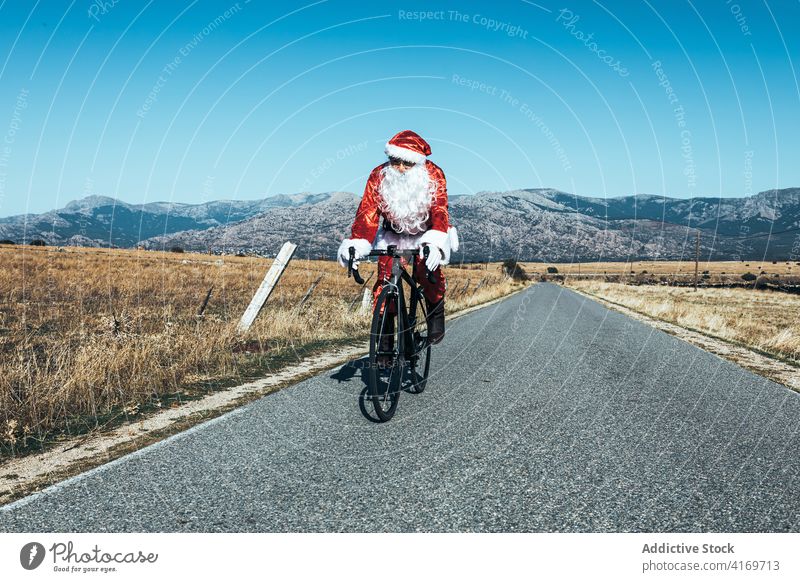Santa Claus riding bicycle along mountain road santa claus ride bike empty costume red color male modern journey freedom speed trip move sunny activity