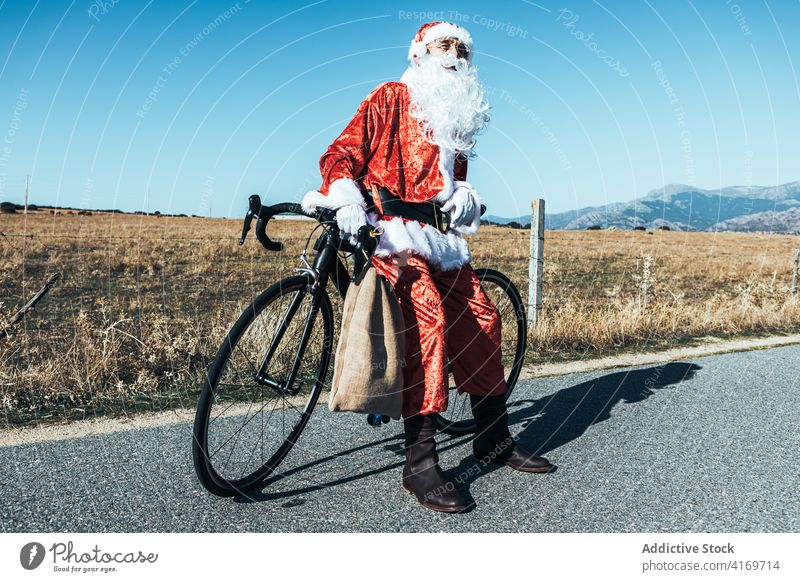 Santa Claus near bicycle on road in countryside santa claus bag relax roadway vehicle bike santa hat male beard costume red color modern style stand transport