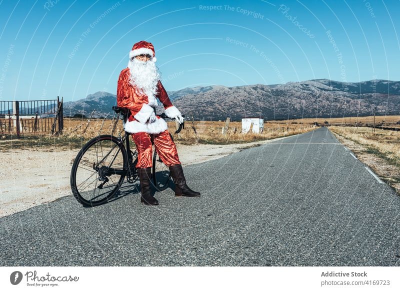 Santa Claus near bicycle on road in countryside santa claus relax roadway vehicle bike santa hat male beard costume red color modern style stand transport