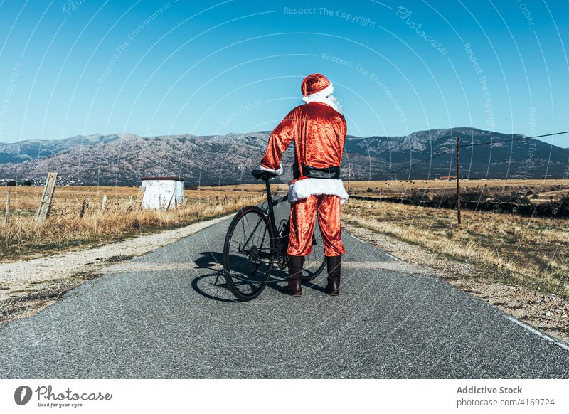 Anonymous Santa Claus near bicycle on road in countryside santa claus relax roadway vehicle bike santa hat male beard costume red color modern style stand