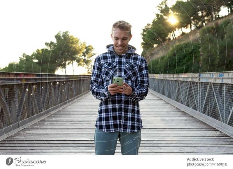Happy man with mobile phone standing on footbridge smartphone using hipster positive check happy traveler young male countryside gadget device lifestyle