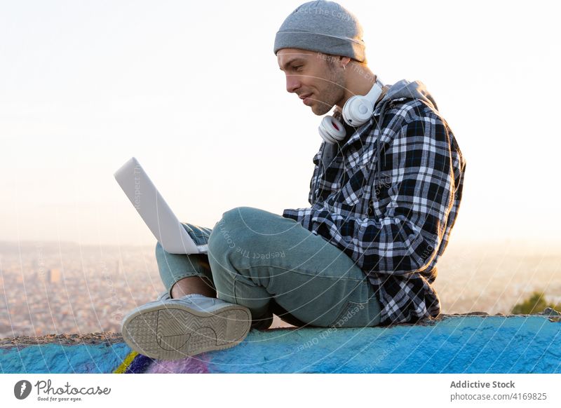 Positive young man in casual wear using laptop on street hipster happy positive browsing online internet blogger freelance sunset device gadget modern lifestyle
