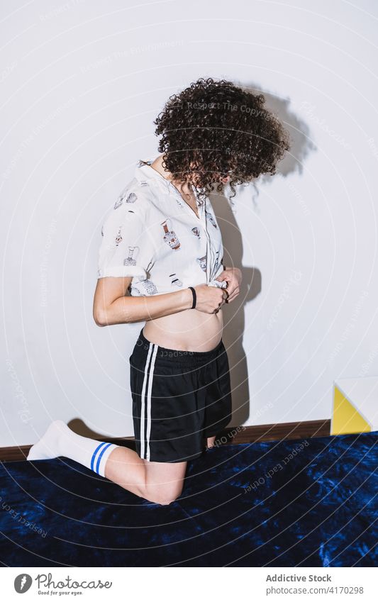 Young woman tying shirt kneeling on bed rest tie tummy cozy curly hair comfort peaceful calm female shorts young slim serene lifestyle casual tranquil bedroom