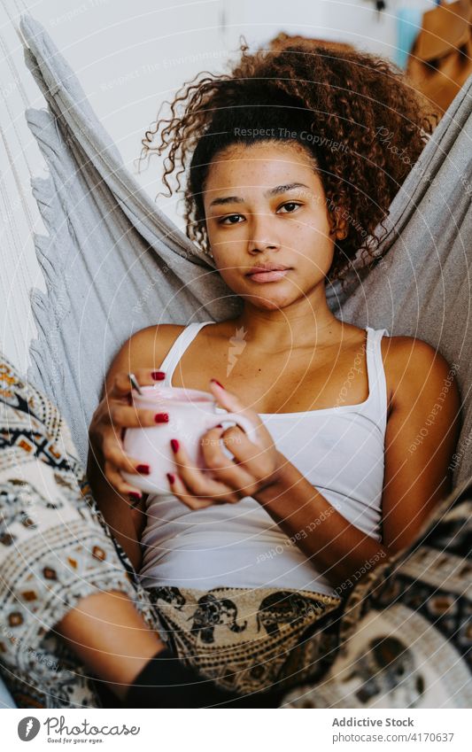 Ethnic woman with cup of coffee resting in hammock at home drink relax chill cozy young female african american black ethnic beverage lifestyle tea weekend