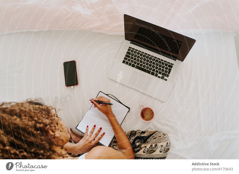 Unrecognizable ethnic woman with laptop and planner working in bedroom at home take note freelance remote casual young female african american black device