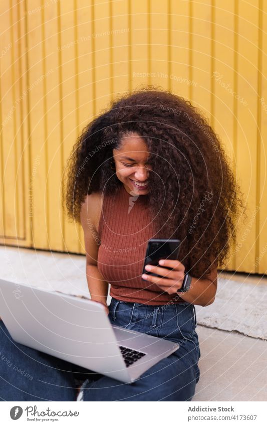 Cheerful African American self employed woman sitting on street with laptop smartphone freelance curly hair city entrepreneur ethnic black african american