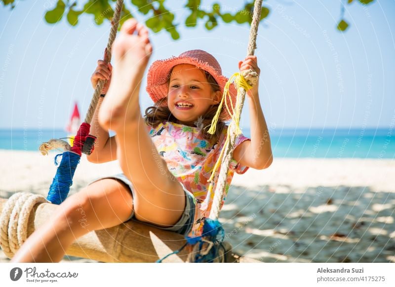 Happy cute little girl laughing and swinging on swing on the tree at the beach. Beautiful summer sunny day, turquoise sea, rocks, white sand, picturesque tropic landscape. Phuket, Thailand. Carefree