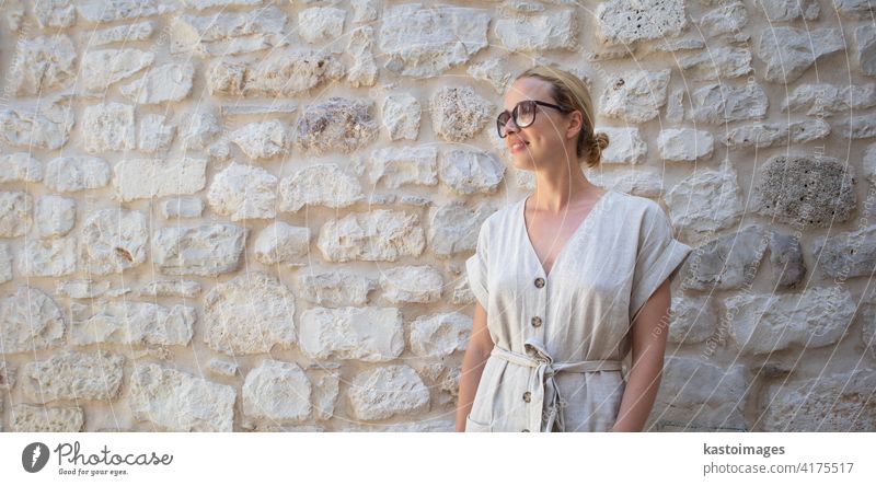 Portrait of beautiful cheerful blonde woman wearing one piece sundress and summer hat, standing in front of old medieval stone wall. Summer vacation portrait concept. Copy space.