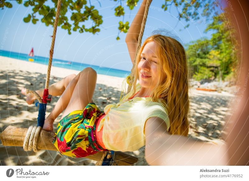 Happy young woman with red hair laughing and swinging on swing on a tree at the beach, taking a selfie. Beautiful summer sunny day, turquoise sea, white sand, tropic landscape. Phuket, Thailand.