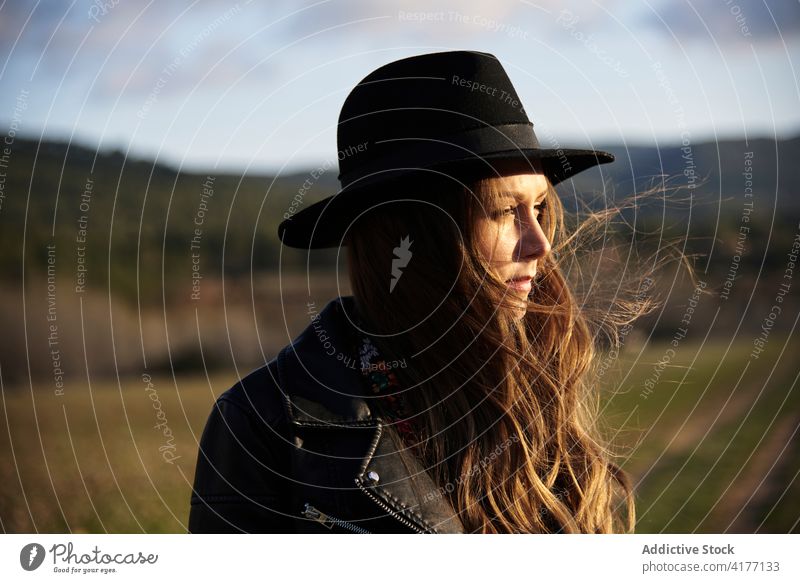 Charming woman in hat and leather jacket in countryside style sunset trendy outfit charming field female black apparel braid hairstyle long hair appearance