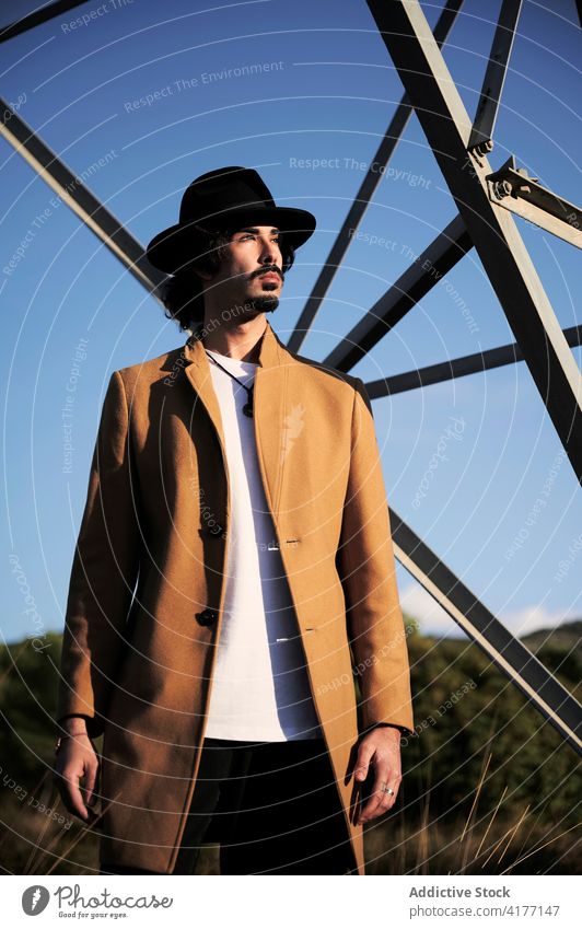 Stylish man in hat and coat standing near metal tower style trendy confident modern serious young beard male ethnic lifestyle outfit personality fashion pensive