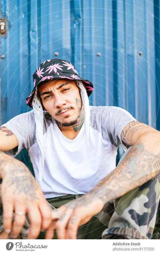 Informal tattooed man sitting near fence informal hipster alternative style break dance subculture young male trendy individuality millennial lifestyle