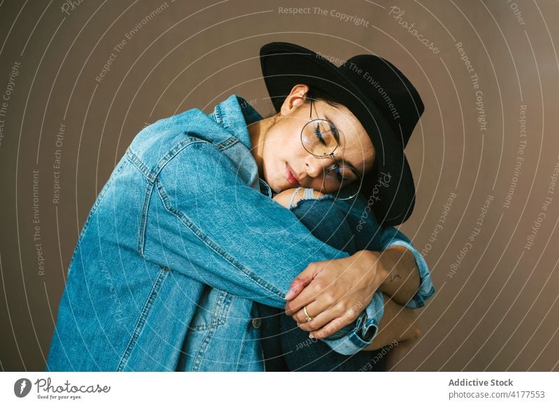 Stylish woman in denim outfit and hat in studio embracing knee trendy style model female stool sit eyes closed calm fashion garment apparel modern young