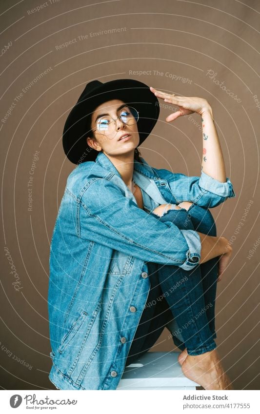 Stylish woman in denim outfit and hat in studio embracing knee touch trendy style model female stool sit calm fashion garment apparel modern young appearance