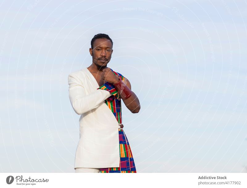 Black man in stylish traditional clothes against sky fashion model style fancy costume apparel extraordinary extravagant serious male ethnic black cloudy blue