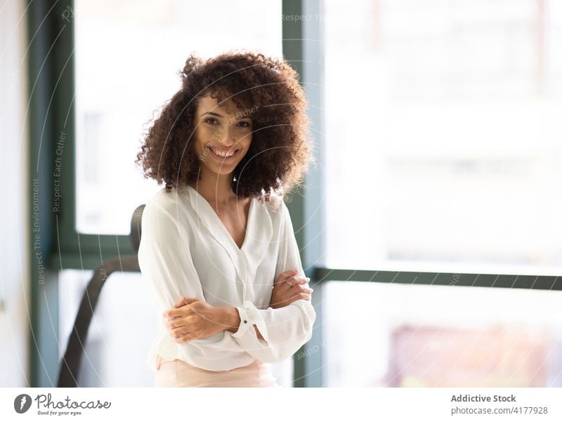 Smiling ethnic woman standing near window in office cheerful smile happy young positive success optimist hispanic female curly hair modern lifestyle glad