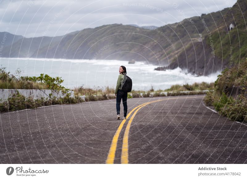 Woman with backpack walking on road on seashore traveler coast rocky woman ocean stormy adventure female active hiker backpacker nature direction route roadway