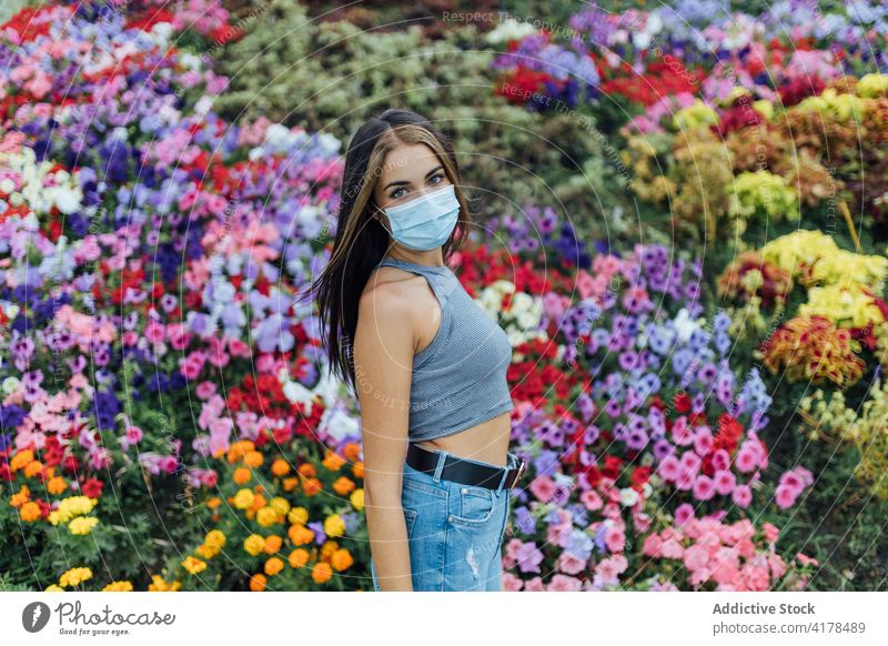 Young woman in medical mask standing near blooming flowers protect park coronavirus covid 19 pandemic new normal young female casual disposable colorful covid19