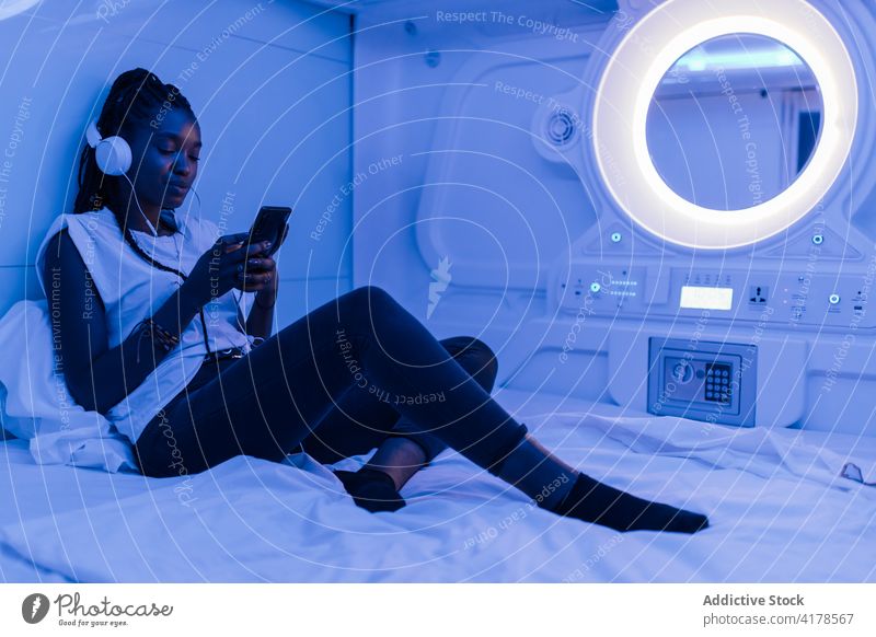 Ethnic woman using smartphone in capsule hotel rest traveler bed headphones browsing mobile listen gadget device online internet surfing connection communicate