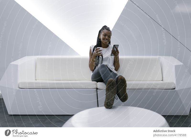 Cheerful black woman resting in white modern living room browsing smartphone futuristic interior geometry furniture cheerful female ethnic african american sofa