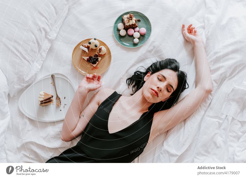 Peaceful woman lying on bed with various desserts hotel luxury serve set sweet assorted female tasty weekend yummy delicious carefree serene eyes closed