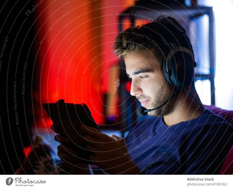 Positive man in headphones browsing on tablet in dark room using darkness sofa content watching gadget rest entertain play cellphone surfing listen device work