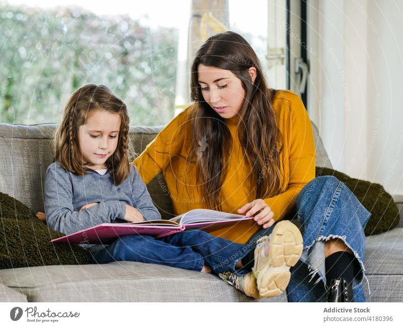 Teenager with sister reading book on sofa in house point spend time interact weekend smile home pastime content girl teen watching show share rest couch cozy