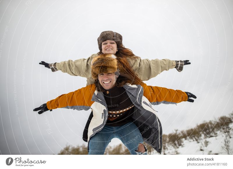 Happy woman riding piggyback on boyfriend during winter trip ride girlfriend arms raised freedom having fun snow mount nature couple lean forward carry