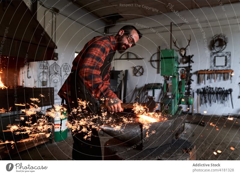 Focused young blacksmith heating and striking metal on anvil in workshop man strike iron smithy spark manual hammer craftsman male beard brutal apron goggles