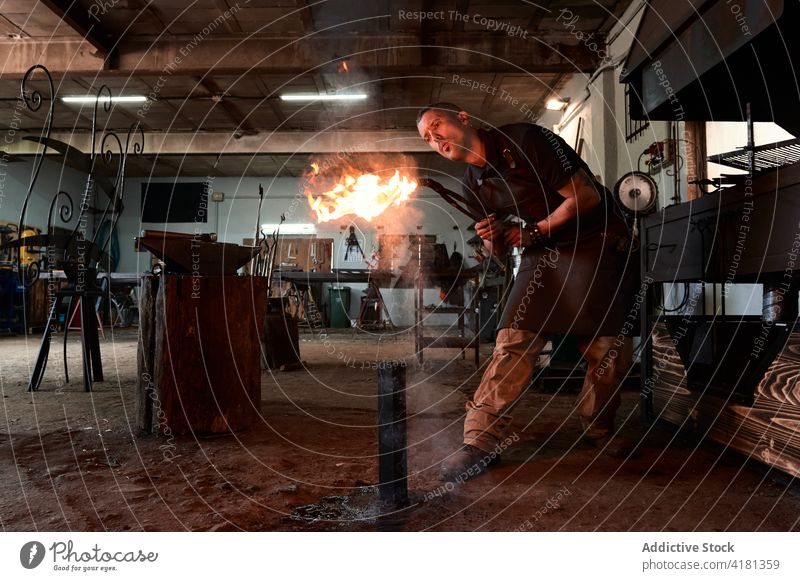 Attentive young blacksmith blowing on fire while heating tongs in workshop man pliers smithy flame concentrate professional occupation skill male apron shabby