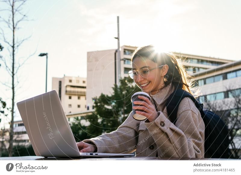 Cheerful female student working on laptop in city park woman toothy smile assignment cheerful using campus gadget break coffee backpack eyeglasses to go