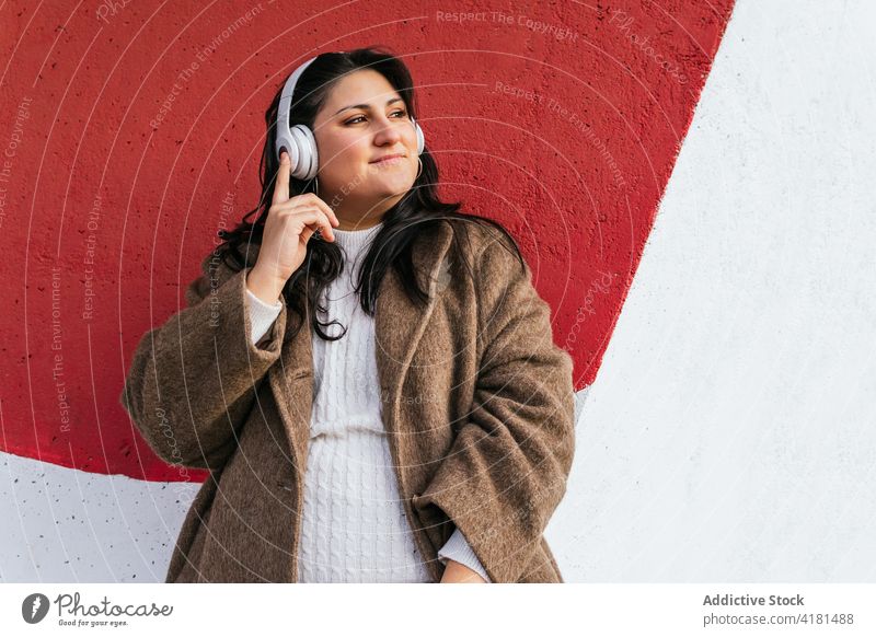 Smiling woman listening to music from headset near bright wall headphones wireless dreamy enjoy meloman street using gadget device admire content idyllic audio