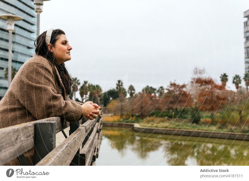 Dreamy woman enjoying pond from fenced embankment in autumn city admire nature dreamy hands clasped reflect idyllic channel water grow vegetate bright building