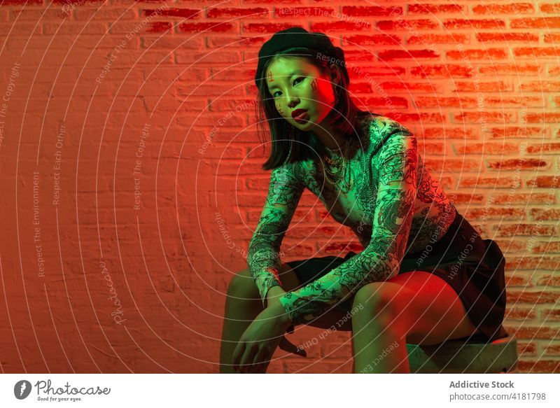 Trendy Asian woman sitting on metal barrel in dark studio style confident grace trendy outfit cool appearance street style brick wall personality hat young