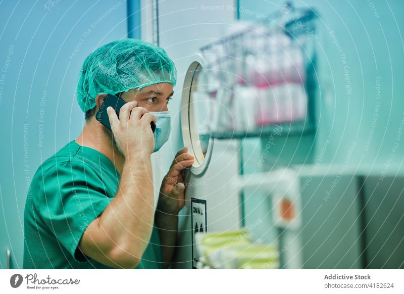 Focused male medic talking on smartphone and looking through window during operation man doctor operating room phone call conversation concentrate medical