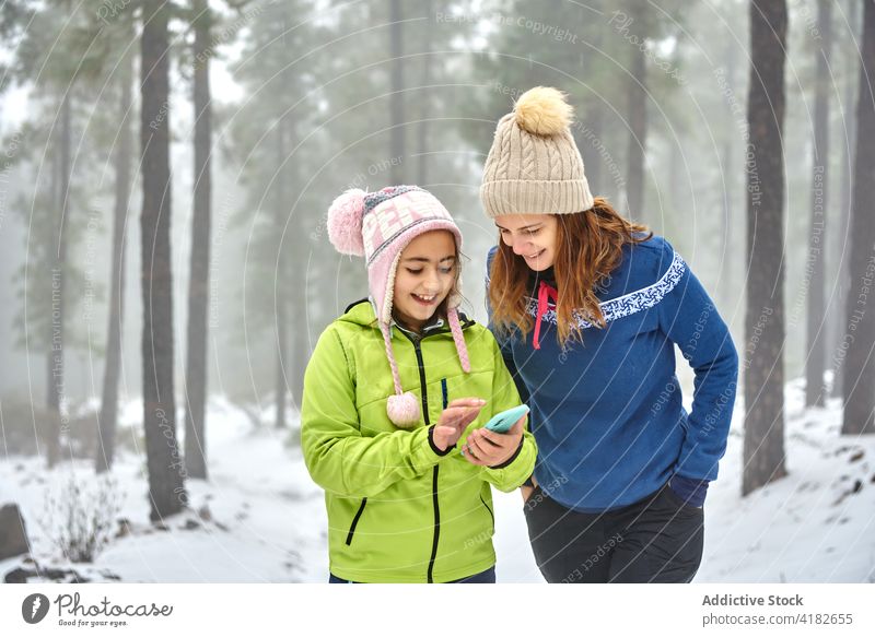 Cheerful girl and mother using smartphone in winter forest woman walk cheerful toothy smile outerwear daughter happy woodland gadget device browsing mobile