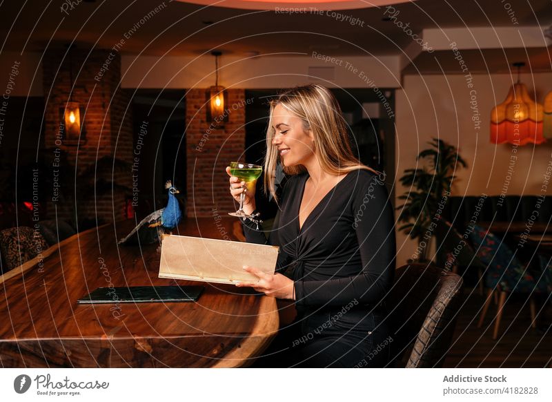 Joyful young woman drinking cocktail and reading menu in stylish restaurant bar smile enjoy positive customer style relax female blond long hair trendy alcohol