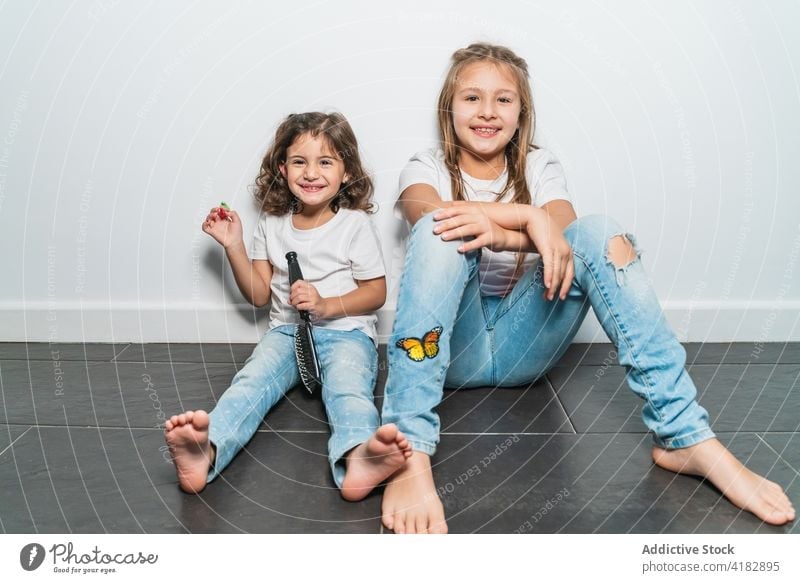 Cheerful little sisters sitting on floor girl cheerful happy cute childhood together joy at home toothy smile enjoy adorable jeans shirt kid wall positive