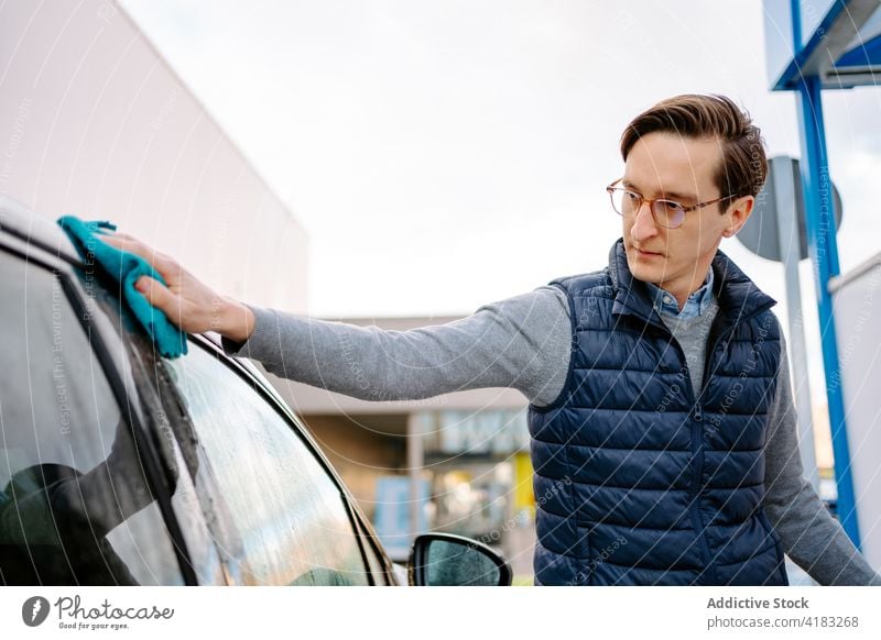 Concentrated young man wiping car with napkin wash wipe rag focus vehicle self service auto station street window water automobile manual male casual eyeglasses