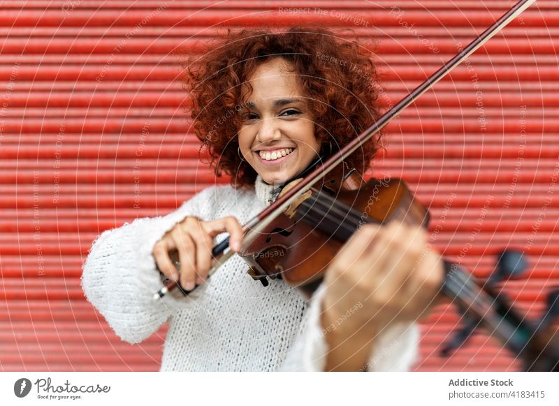 Cheerful redhead woman playing violin against red wall musician happy toothy smile instrument acoustic perform live melody talent skill attractive sound song