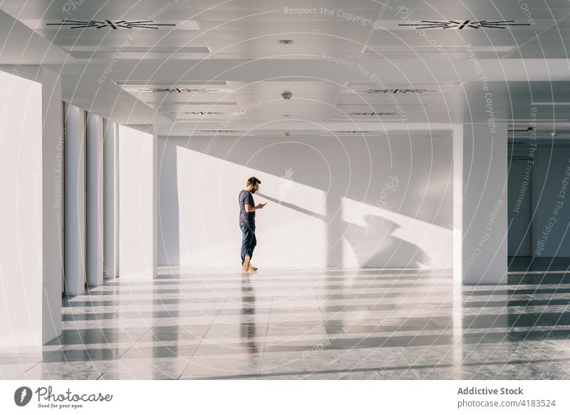 Man standing in empty spacious office hall and browsing smartphone man facility shadow using male interior sunlight geometry shape hallway white wall surfing