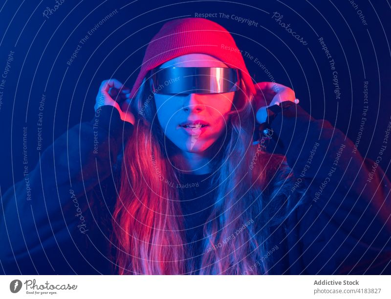 Stylish young lady in trendy sunglasses standing in dark room with neon lights woman style self assured appearance fashion futuristic informal portrait