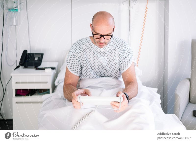 Focused male patient using tablet on ward bed man focus workaholic device hospital clinic watch professional concentrate digital medicine sick ill eyeglasses