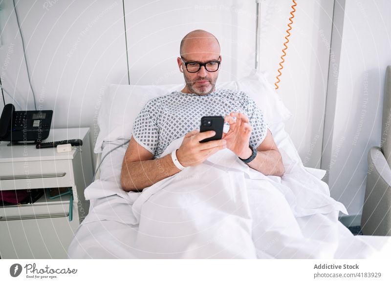Focused male patient browsing phone on ward bed man smartphone using mobile focus workaholic device hospital clinic watch professional concentrate digital